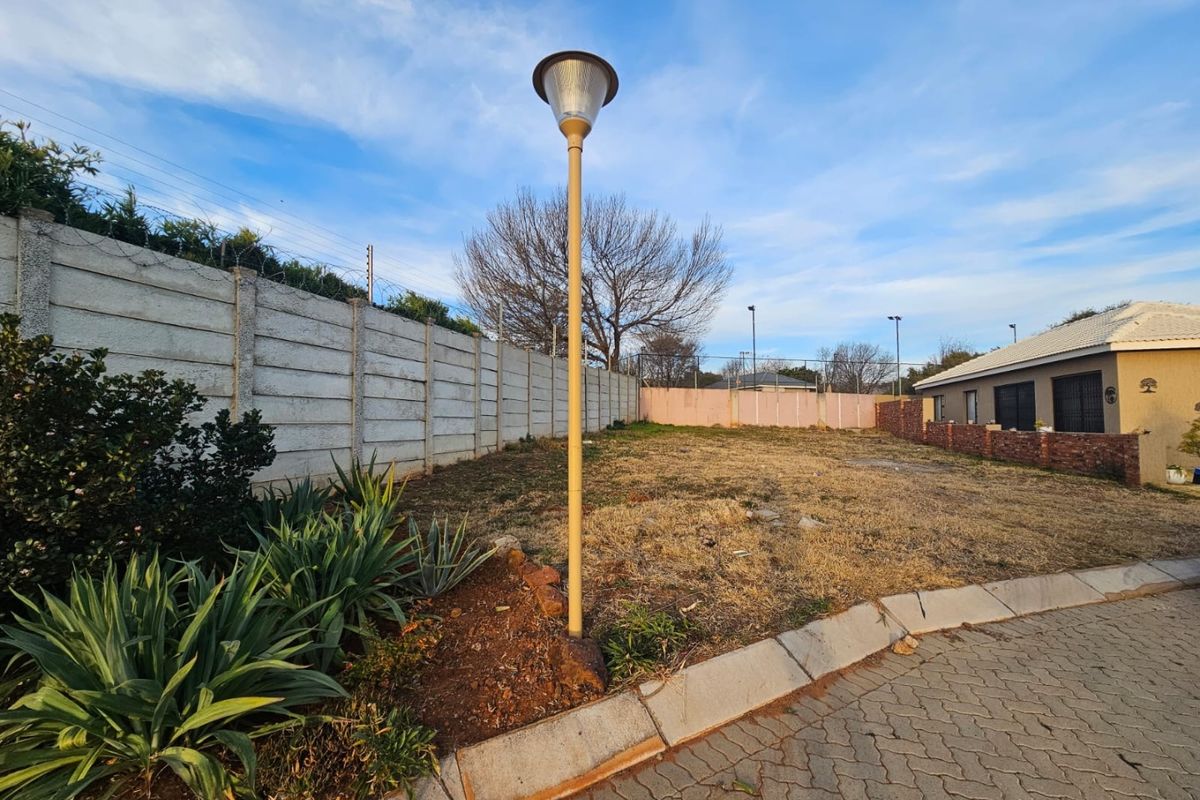 A vacant residential plot featuring a precast concrete boundary wall with security fencing, paved driveway access, and an existing street lamp.
