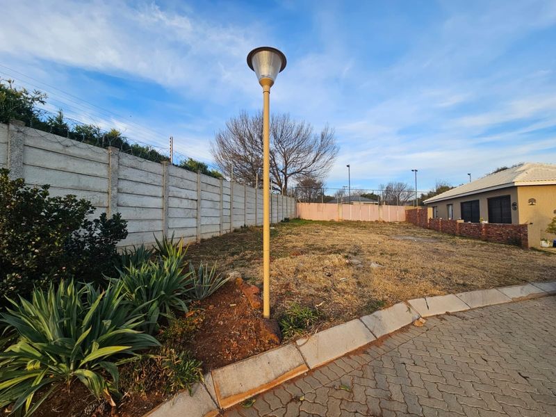 A vacant residential plot featuring a precast concrete boundary wall with security fencing, paved driveway access, and an existing street lamp.