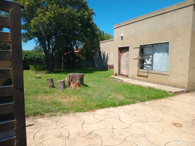 Rear exterior view of the property featuring a paved patio area, lawn, and louvered windows with visible wear to the exterior wall and door frame.