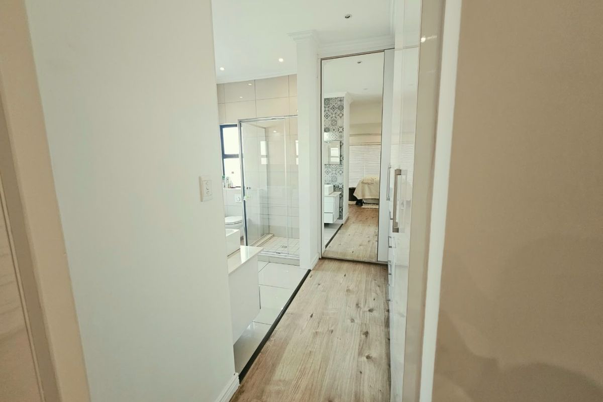 A modern bathroom featuring a glass shower enclosure, built-in vanity, and mirrored wardrobe doors, connecting to a well-lit dressing area with wood-look flooring.
