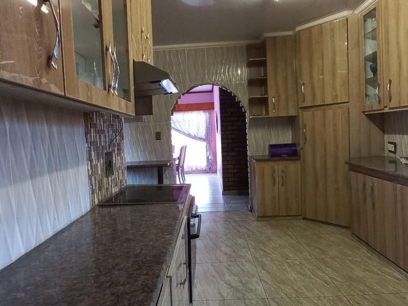 A functional kitchen featuring wood-look laminate cabinetry, stone-effect countertops, tiled backsplash, and an integrated hob with extractor fan, leading through an archway.