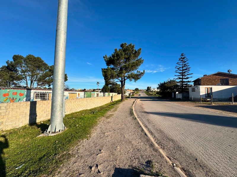 A street view showing a paved road, grass verge, and a concrete block boundary wall in a residential neighborhood.