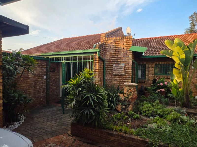 Front entrance view featuring a face brick facade, tiled roof, paved driveway, and a well-established garden with a raised planter bed.
