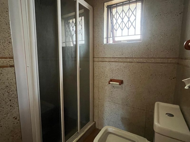 A functional bathroom featuring a shower enclosure, toilet, and tiled finishes with a small window fitted with security bars.