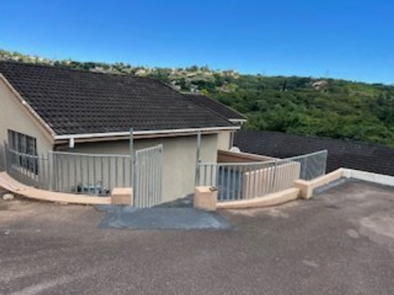Front exterior view of the property featuring a tiled roof, paved driveway, and metal security fencing with a hillside backdrop.