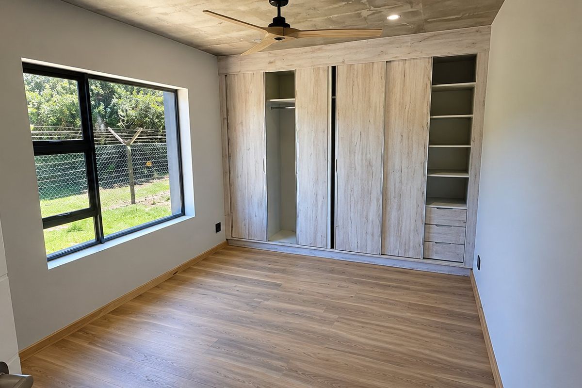 A modern, empty bedroom featuring laminate flooring, an exposed concrete ceiling, a ceiling fan, and extensive built-in wooden cupboards.