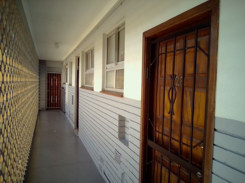 An exterior apartment hallway featuring a timber entrance door with a security gate, aluminium windows, and a decorative privacy screen wall.