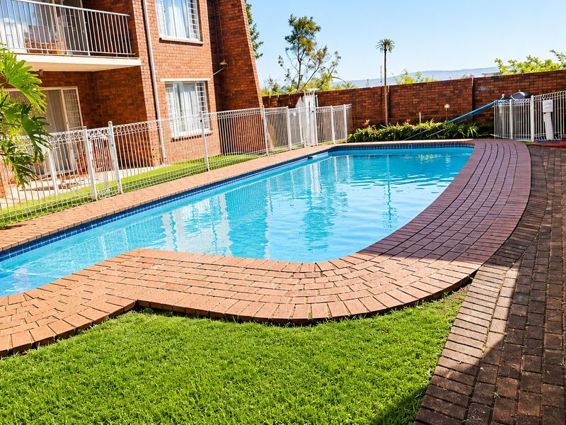 A well-maintained communal swimming pool area featuring brick paving, secure metal fencing, and a landscaped lawn, set within an apartment complex.