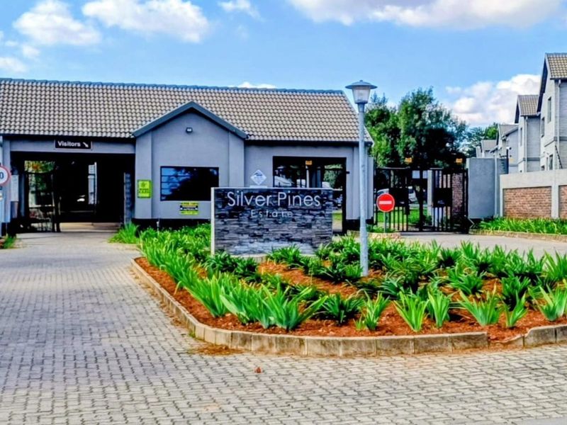 The entrance to Silver Pines Estate featuring a paved driveway, security gatehouse, and landscaped garden beds.