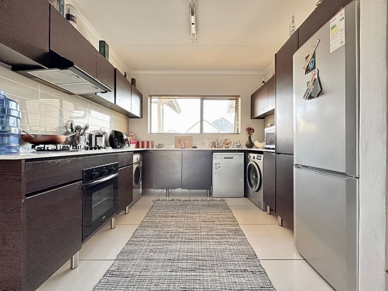 A functional galley-style kitchen featuring dark wood cabinetry, integrated appliances, tiled splashbacks, and light-colored floor tiles.