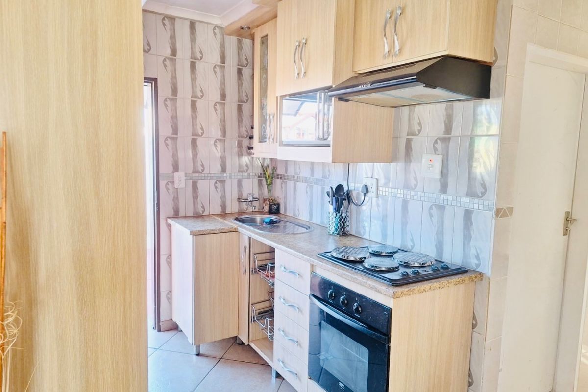 A compact kitchen featuring light wood-finish cabinetry, tiled backsplash, built-in oven, electric hob, and extractor fan with tiled flooring.
