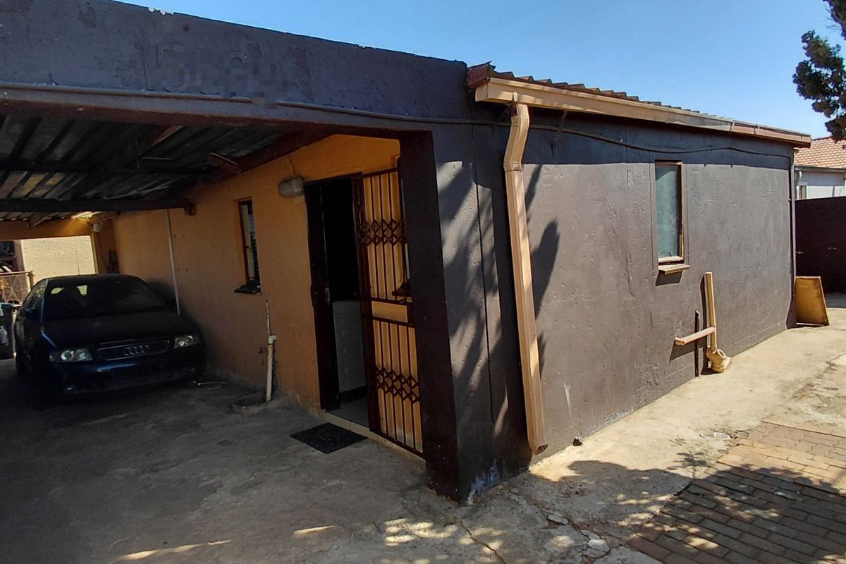 Rear exterior view of the property featuring a covered carport, security gate, and paved driveway area with visible wear to the exterior finishes.