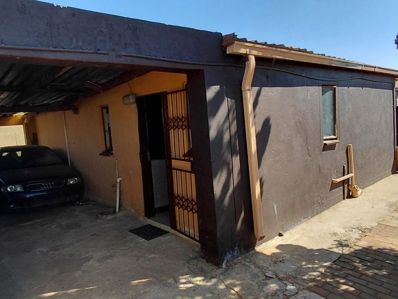 Rear exterior view of the property featuring a covered carport, security gate, and paved driveway area with visible wear to the exterior finishes.