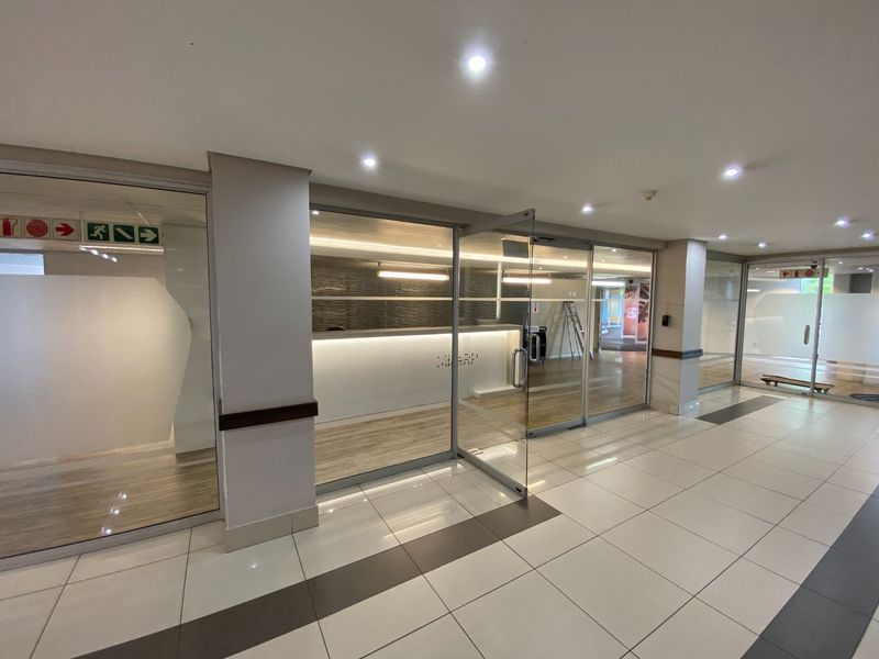 Modern commercial entrance area featuring glass partitioning, a built-in reception desk, and clean tiled flooring with recessed lighting.