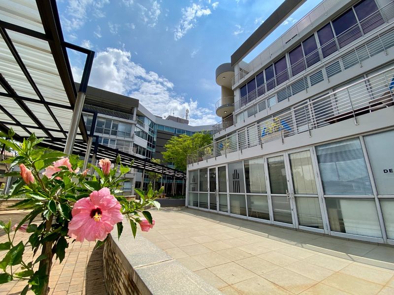 A modern commercial building exterior featuring a paved terrace, metal pergola structure, and multi-story glass facade with stainless steel balcony railings.