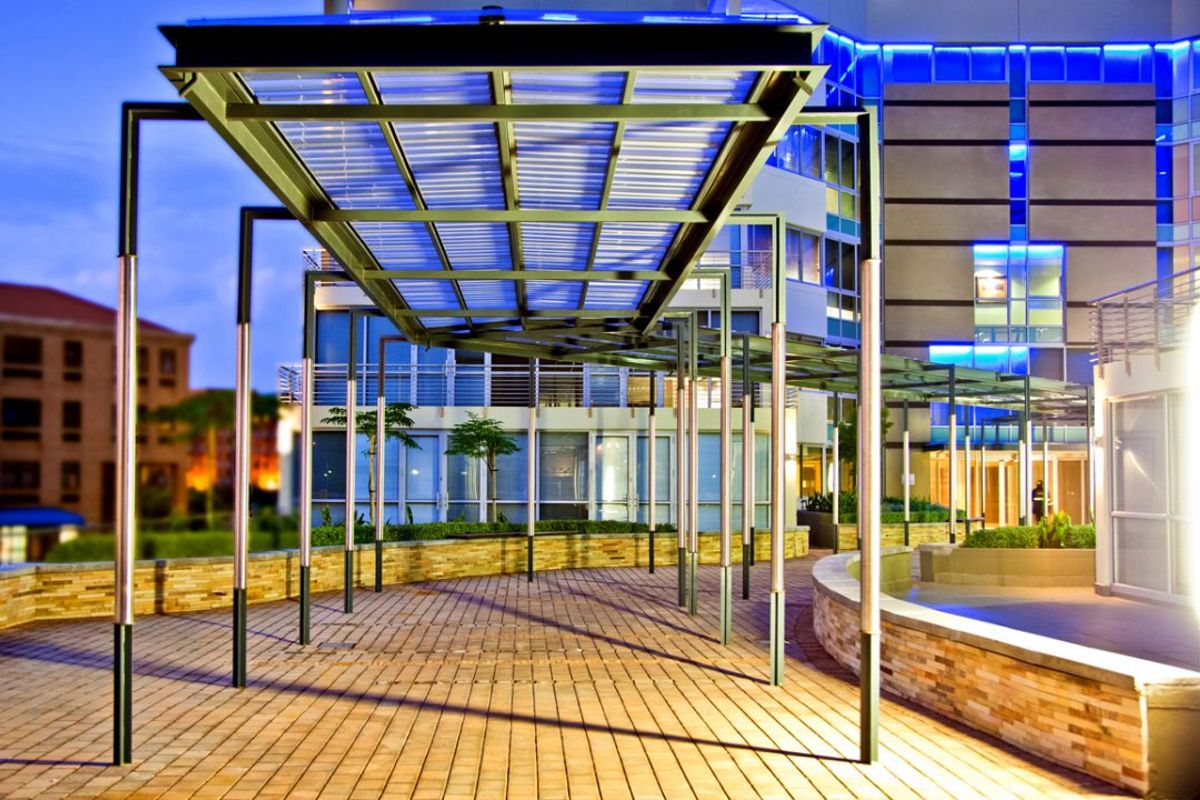 A modern commercial courtyard featuring a covered metal walkway, stone-clad retaining walls, and integrated architectural facade lighting.
