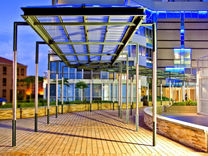 A modern commercial courtyard featuring a covered metal walkway, stone-clad retaining walls, and integrated architectural facade lighting.