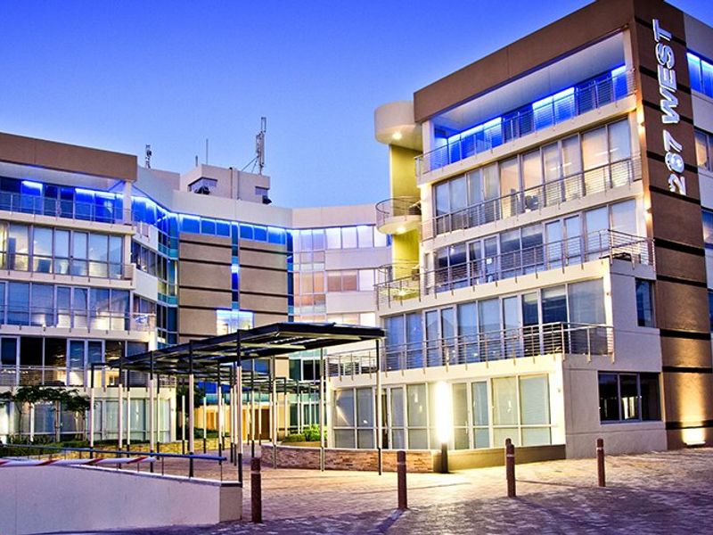 Modern multi-story commercial office building featuring a glass facade, integrated balcony levels, and a paved entrance area with secure access.