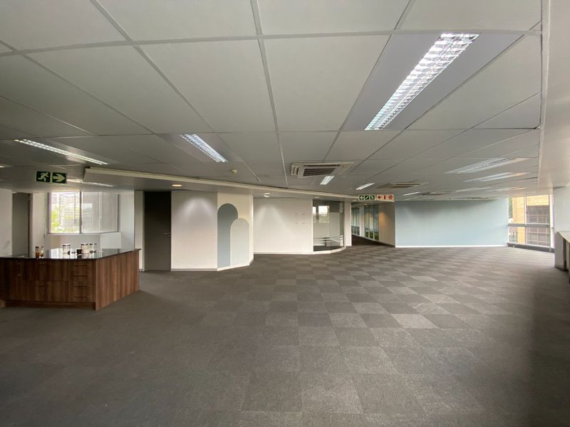 Spacious open-plan commercial office area featuring carpeted flooring, suspended ceilings with recessed lighting, integrated air conditioning, and a small built-in kitchenette unit.