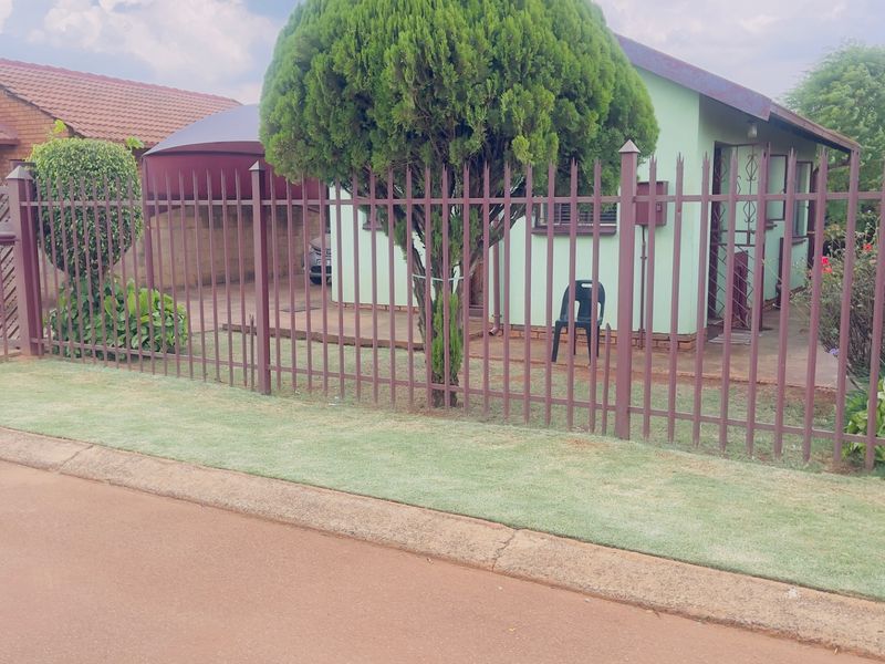 Front view of a residential house featuring a metal security fence, paved driveway, and a carport.