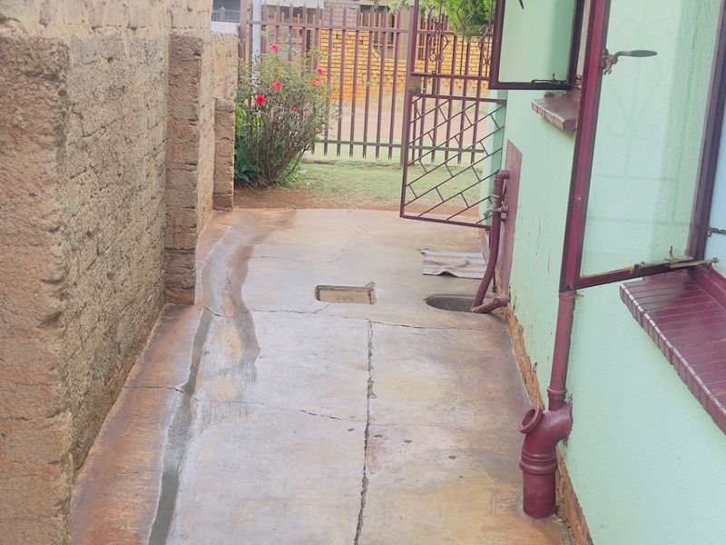 A narrow rear exterior walkway featuring concrete paving, a brick boundary wall, and metal security gate access.