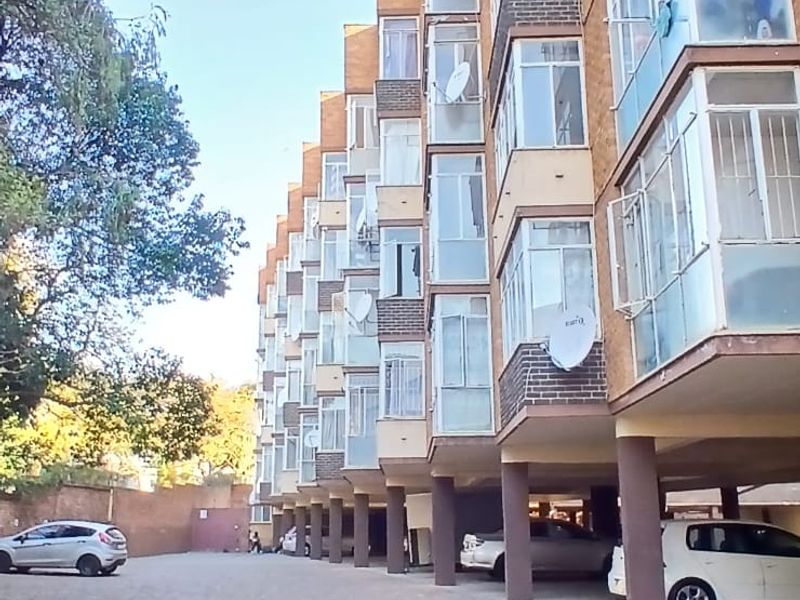Exterior view of a multi-story residential apartment building featuring brick facade, enclosed balconies, and a paved ground-level covered parking area.