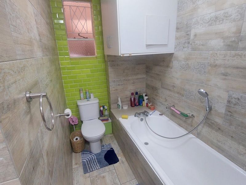 A compact bathroom featuring a built-in bathtub with shower mixer, a toilet, and wall-mounted storage cabinetry, finished with modern wood-look and green accent tiling.