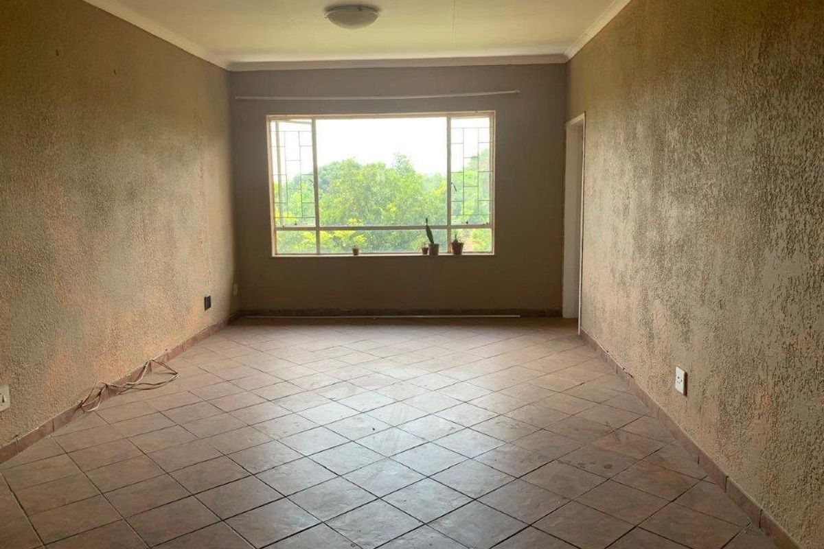 A spacious, empty living area featuring tiled flooring, textured walls, and a large window with security bars, offering a blank canvas for renovation.