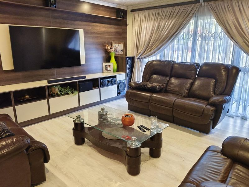 A well-maintained living room featuring a modern built-in media wall unit, light-toned laminate flooring, and large windows with curtains and security gates.
