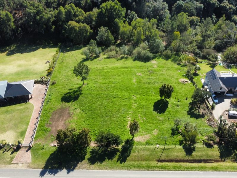 An aerial view of a vacant residential plot featuring established trees, grassed terrain, and perimeter fencing, situated between existing residential properties.