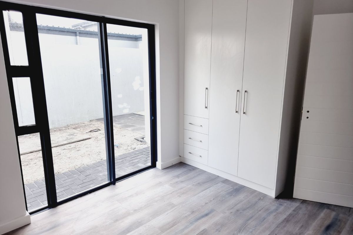 A bright, modern bedroom featuring built-in cupboards, light-toned laminate flooring, and a large sliding glass door providing direct access to the outdoor area.