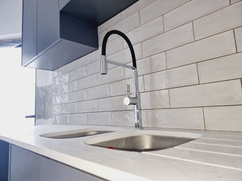 Modern kitchen area featuring a double stainless steel sink, gooseneck faucet, stone countertop, and a stylish subway tile backsplash with dark cabinetry.