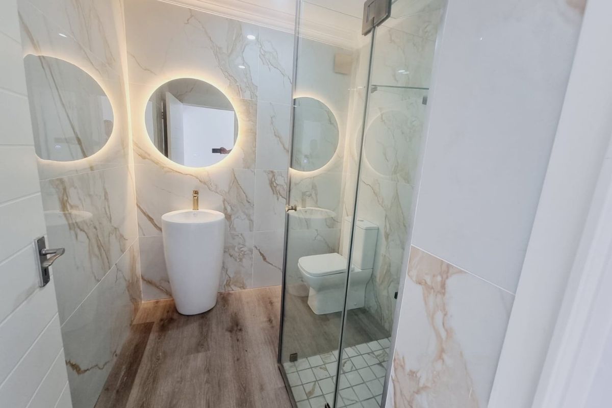 Modern bathroom featuring marble-look wall tiles, wood-look flooring, a pedestal basin with gold faucet, backlit circular mirror, and a glass-enclosed shower.