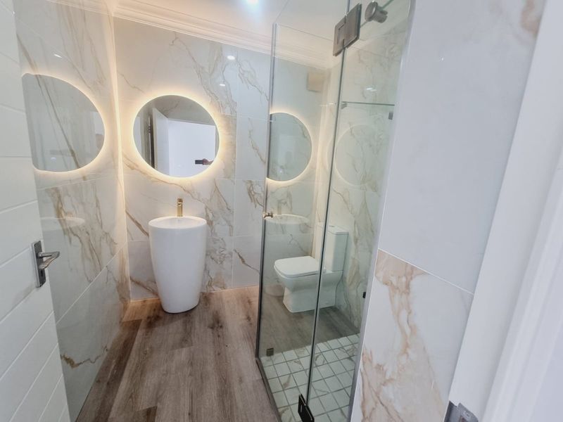Modern bathroom featuring marble-look wall tiles, wood-look flooring, a pedestal basin with gold faucet, backlit circular mirror, and a glass-enclosed shower.
