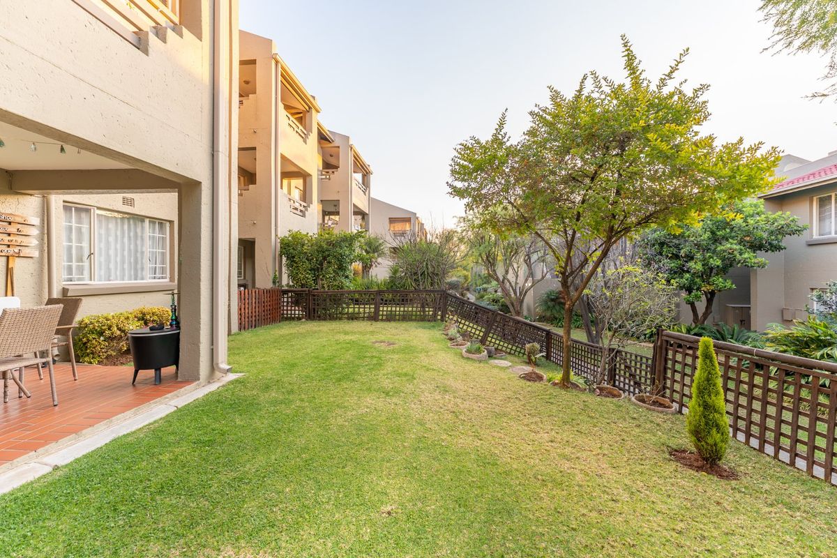 A well-maintained rear garden featuring a covered patio with tiled flooring, established trees, and timber fencing within a townhouse complex.