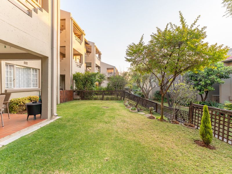 A well-maintained rear garden featuring a covered patio with tiled flooring, established trees, and timber fencing within a townhouse complex.