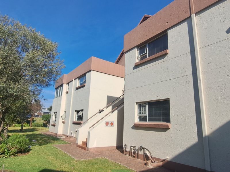 Rear view of a townhouse complex featuring a paved walkway, external staircase, and established garden area with security bars on windows.