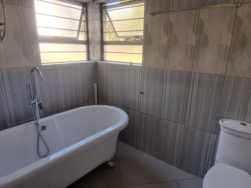 A functional bathroom featuring a freestanding bathtub, toilet, and neutral wall and floor tiling with natural light from top-hung windows.