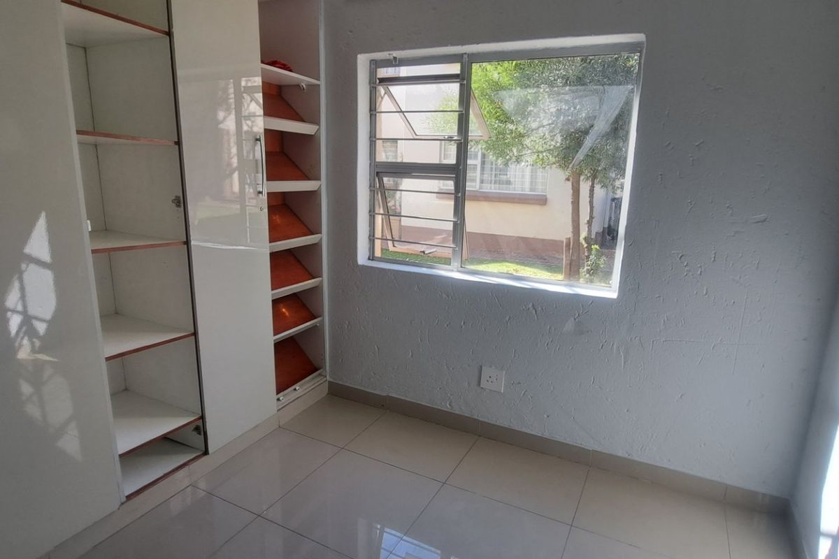 A bright, empty bedroom featuring modern white built-in cupboards, neutral tiled flooring, and a window overlooking the exterior grounds.
