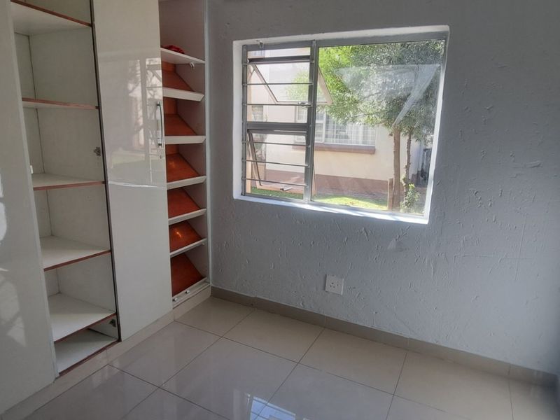 A bright, empty bedroom featuring modern white built-in cupboards, neutral tiled flooring, and a window overlooking the exterior grounds.