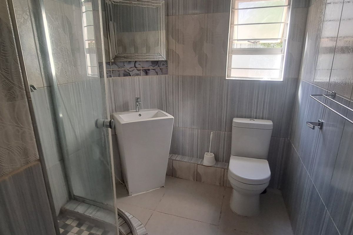 A functional bathroom featuring a shower enclosure, pedestal basin, toilet, and neutral wall tiling with a window for natural light.