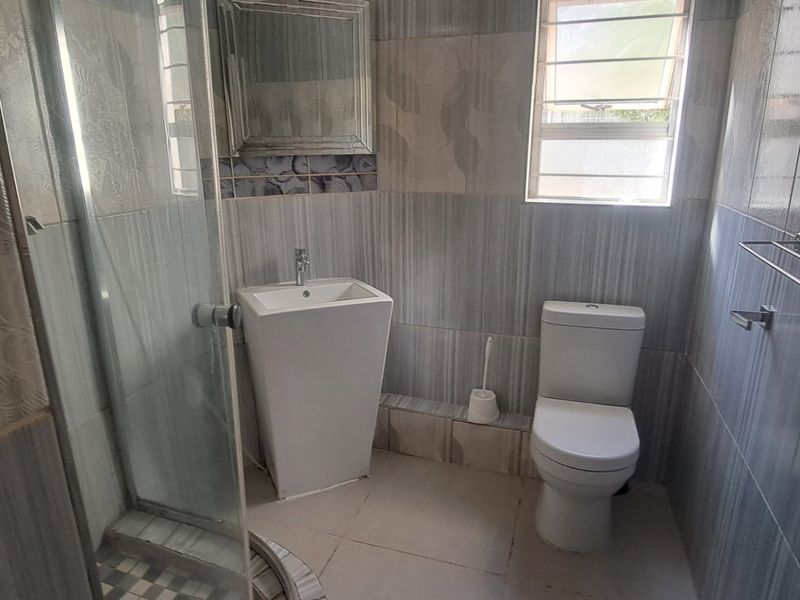 A functional bathroom featuring a shower enclosure, pedestal basin, toilet, and neutral wall tiling with a window for natural light.