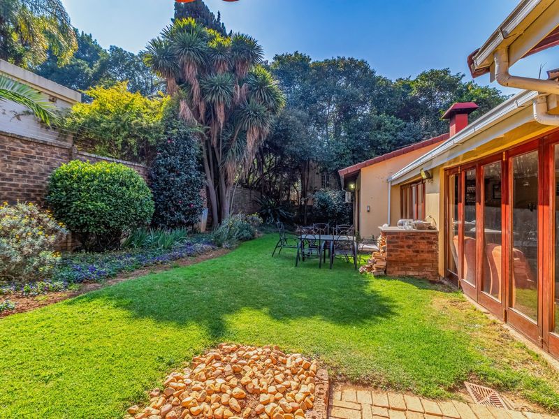 A well-maintained rear garden featuring a lush lawn, established trees, a brick boundary wall, and a paved patio area with a built-in brick braai.