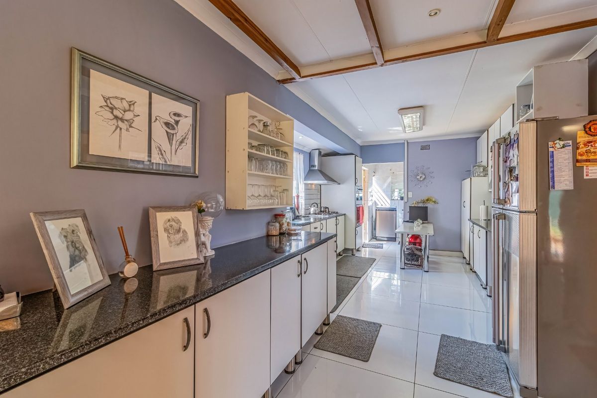 A functional kitchen featuring granite countertops, ample built-in cabinetry, tiled flooring, and exposed ceiling beams, leading through to a secondary utility area.
