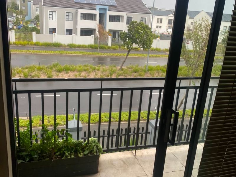 A tiled balcony featuring a metal balustrade, accessible via a sliding glass door, overlooking a street and neighboring residential buildings.