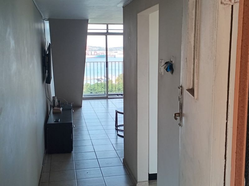 A narrow hallway leading towards a living area with sliding glass doors and balcony access, featuring tiled flooring and neutral wall finishes.