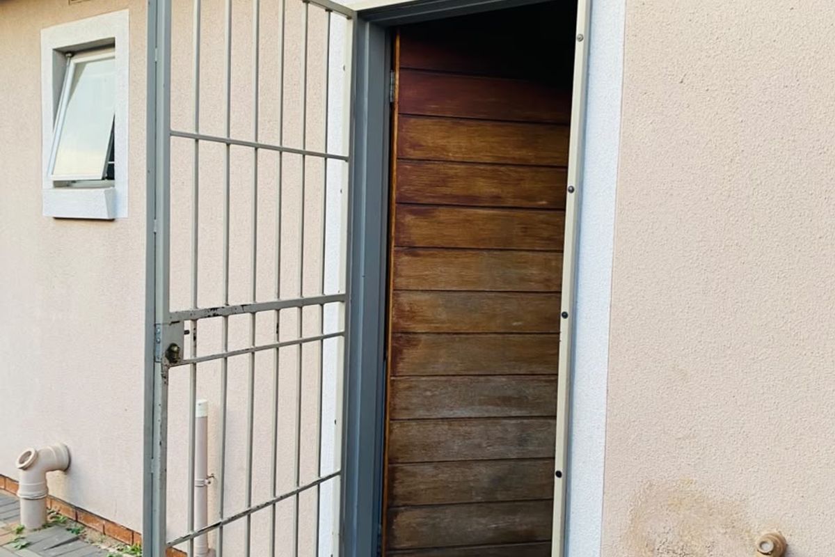 Rear exterior view showing a timber door with a security gate, paved walkway, and external plumbing fixtures.