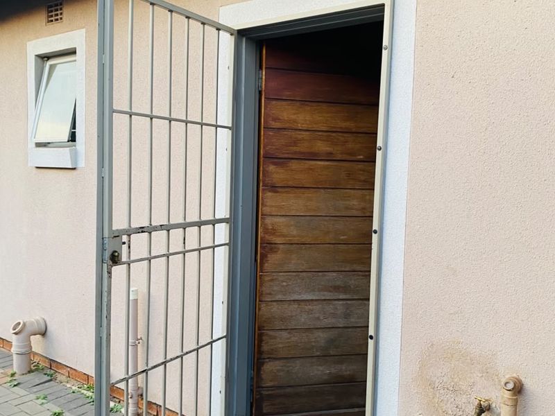 Rear exterior view showing a timber door with a security gate, paved walkway, and external plumbing fixtures.