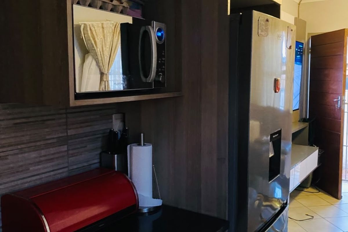 A compact kitchen area featuring modern wood-finish cabinetry, dark countertops, a built-in microwave, and tiled flooring, leading into an adjacent living space.