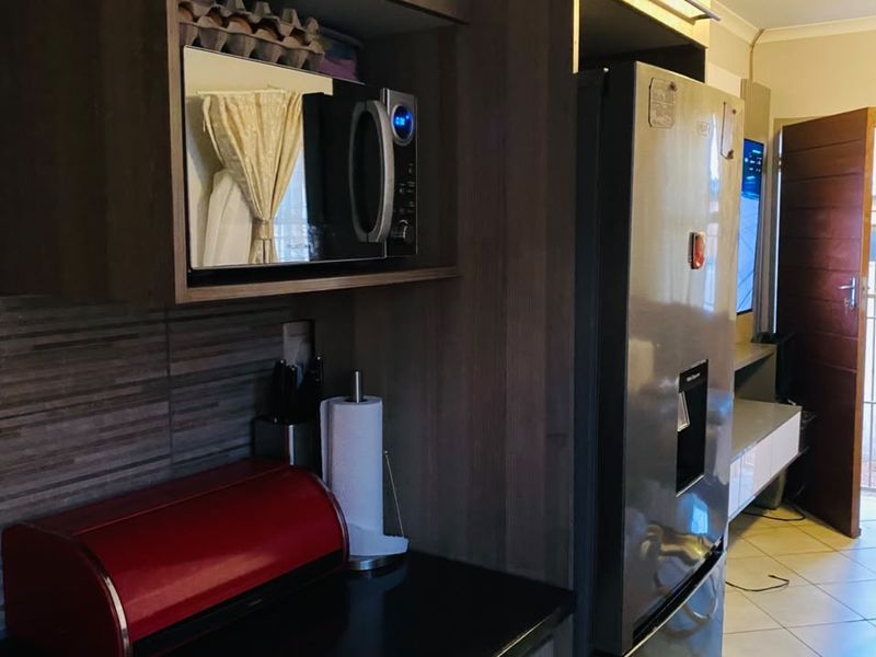 A compact kitchen area featuring modern wood-finish cabinetry, dark countertops, a built-in microwave, and tiled flooring, leading into an adjacent living space.